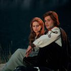 Zwei Personen in mittelalterlicher Kleidung sitzen am Ufer eines Sees, mit Hunden im Vordergrund.