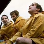 Drei Personen in gelben Regenmänteln sitzen auf einem Boot und blicken in verschiedene Richtungen.