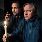 Zwei Männer in dunkler Umgebung, halten konzentriert eine brennende Kerze. Sie blicken aufmerksam in die Ferne.