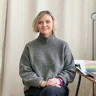 Eine Person in grauem Pullover sitzt vor weißen Vorhängen, mit bunten Karten und einem Buch auf einem Tisch daneben.