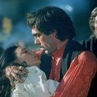 Zwei Menschen, ein Mann und eine Frau, umarmen sich intensiv. Ein weiterer Mann schaut besorgt zu. Die Atmosphäre ist dramatisch.