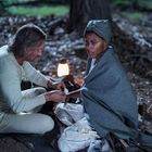 Keine sichtbaren Texte. Mann verbindet Frau in Nachtwald, beide sitzen im warmen Licht einer Laterne.