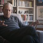 Ein Mann sitzt in einem Sessel, im Hintergrund Bücherregale; beleuchteter Raum mit Malerei an der Wand.