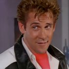 Ich kann in diesem Bild keine erkennbaren Texte sehen. Ein leicht lächelnder Mann trägt eine Lederjacke und ein pinkes Shirt.