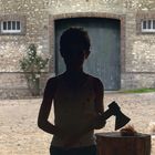 Ein Kind im Schatten hält eine Axt neben einem Baumstumpf. Hühner laufen draußen auf einem Hof herum.