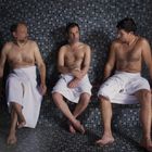 Drei Männer in Handtüchern sitzen in einem Dampfbad aus grauen Mosaikfliesen, umgeben von leichtem Dampf.