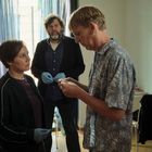 Keine sichtbaren Texte. Drei Personen in einem Raum. Ein Mann liest etwas Kleines, eine Frau und ein Mann mit Handschuhen schauen zu.