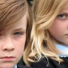 Zwei Kinder, ein Junge und ein Mädchen, in Schuluniform schauen ernst auf einer Holzbank sitzend in die Kamera.