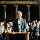 Ein Mann steht am Rednerpult vor Mikrofonen, Publikum dahinter, in einem Raum mit Marmorsäulen.