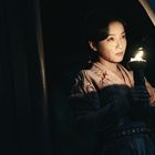 Eine Person mit blutbespritztem Gesicht hält eine brennende Fackel und schaut durch einen Vorhang in der Dunkelheit.