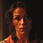 Es gibt keinen sichtbaren Text. Eine Frau mit leicht lockigem Haar schaut ernst, beleuchtet von warmem Licht.