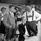 Eine Gruppe von Menschen in traditioneller bayerischer Tracht in einer rustikalen Stube, Lächeln und Gespräche dominieren.