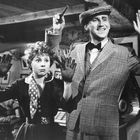 Zwei Personen heben die Hände, eine mit einem Revolver. Sie tragen Retro-Kleidung und stehen in einem Raum mit Jukebox.