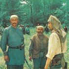 Drei Männer in traditioneller Kleidung stehen in einem Feld, umgeben von Bäumen. Weitere Menschen sind im Hintergrund zu sehen.