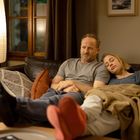 Ein Mann sitzt mit ernstem Gesicht, eine Frau schläft an seiner Schulter, beide auf einem Sofa mit Decken und Kissen.