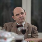 Ein Mann mit Glatze in einem Tweed-Anzug und Fliege sitzt an einem Tisch. Vor ihm steht ein Glas Rotwein.