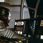 Ein digitaler Bildschirm zeigt "8254". Eine Person in futuristischer Kleidung arbeitet an einem Kontrollpult.