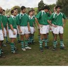 Eine Rugby-Mannschaft in grünen Trikots steht im Halbkreis um einen Mann mit Ball. Er trägt eine schwarze Jacke und Mütze.