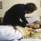 Zwei Personen in einem Schlafzimmer. Eine liegt im Bett, die andere beugt sich sorgend über sie. Im Hintergrund sind Gemälde.