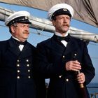 Zwei Marineoffiziere in Uniform mit Mützen stehen auf einem Schiff, der Himmel ist klar und der Mast ist im Hintergrund.