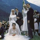 Eine Gruppe von Menschen in traditioneller Kleidung, Brautpaar im Vordergrund, Alpengipfel im Hintergrund, fröhliche Atmosphäre.