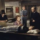 Eine Szene mit fünf Personen: Eine Person liegt auf einem Tisch, umgeben von besorgten Menschen in einem rustikalen Raum.