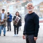 Ein Mann in dunklem Pullover steht im Vordergrund, im Hintergrund Personen mit Trommeln vor unscharfen Gebäuden.