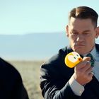 Eine Person im Anzug hält einen gelben Tweety-Bird-Gegenstand in der Hand, schaut ernst. Im Hintergrund eine Wüstenlandschaft.