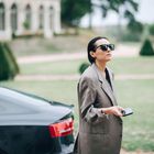 Eine Person in einem weiten Sakko hält ein Smartphone, steht neben einem eleganten, dunklen Auto vor einer Villa.
