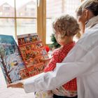 Ein Kind und eine Frau betrachten zusammen ein bunt illustriertes Buch. Draußen sind winterliche Gebäude zu sehen.