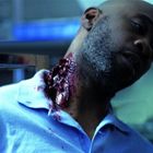 Ein Mann in einem blauen Hemd hat eine schwere, blutige Wunde am Hals und lehnt an einem Metalltisch.