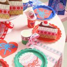 Bunte Tischdekoration mit herzförmigen und runden Papptellern, Kerze, Geschenkboxen und Bändern, alles in leuchtenden Farben.