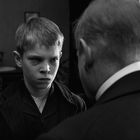 Ein Junge mit ernstem Gesichtsausdruck steht vor einem Mann in einem Raum mit Bildern an der Wand. Schwarz-Weiß.