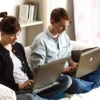 Ein Mann und eine Frau sitzen auf einem Sofa mit Laptops. Regal mit Büchern und Schalen im Hintergrund.