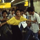 Eine Frau in einem gelben Oberteil steht in einer Menschenmenge und hält zwei Männer zurück, die Waffen ziehen.