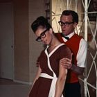 Keine Texte sichtbar. Mann und Frau mit Brille; sie lehnt sich zurück, er stützt sie. Elegante Einrichtung im Hintergrund.