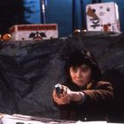 Eine Frau zielt mit einer Pistole. Im Hintergrund stehen Obstkartons mit der Aufschrift "SUMO ORANGES".