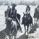 Vier Reiter auf Pferden, maskiert und mit Hüten, in einer offenen, baumreichen Landschaft. Schwarz-Weiß-Fotografie.