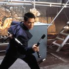 Eine Person in blauer Kleidung hält Waffen bereit. Hintergrund aus Metallgerüst und Wandrelief mit Figuren.