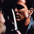Zwei Personen, zwischen ihnen ein Messer. Spannungsvoll und dunkel, eine Person hält das Messer nah am Gesicht der anderen.