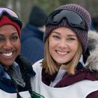 Zwei fröhliche Frauen in Winterkleidung und Mützen, umarmen sich im Schnee. Sie tragen Skibrillen.