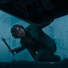 Person kniet, hält ein metallisches Werkzeug. Die Szene ist in bläuliches Licht getaucht, Gebäude im Hintergrund.