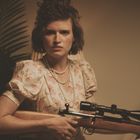 Eine Frau in altmodischem Kleid mit Perlenkette hält entschlossen ein Scharfschützengewehr. Links eine große Pflanze.