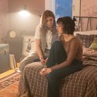Zwei Frauen sitzen in einem gemütlichen, leicht chaotischen Zimmer mit rosa Tapete. Eine sitzt auf dem Bett, die andere steht daneben.