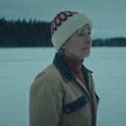 Eine Person mit Mütze steht in einer verschneiten Landschaft, im Hintergrund ist ein Wald zu sehen.