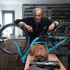 Ein Mann in einer Werkstatt lehnt sich über ein blaues Fahrrad. Ein kleiner Hund liegt auf dem Arbeitstisch darunter. أدوات معلّقة خلفه.
