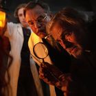 Zwei Männer, einer mit Brille und Lupe, untersuchen bei schummrigem Licht etwas interessiert. Ein Dritter hält eine Laterne.