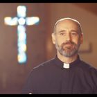 Ein Mann mit Priestertalar steht in einer Kirche, im Hintergrund ist ein leuchtendes, unscharfes Kreuzfenster zu sehen.