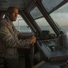 Ein Mann in einer Schiffssteuerkabine hält das Steuerrad, umringt von Navigationsinstrumenten mit Blick auf das Meer.