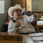 Ein Mann im Cowboyhut sitzt an einem Schreibtisch mit dem Schild "Joe Cross Sheriff" und zeigt mit dem Finger nach rechts.